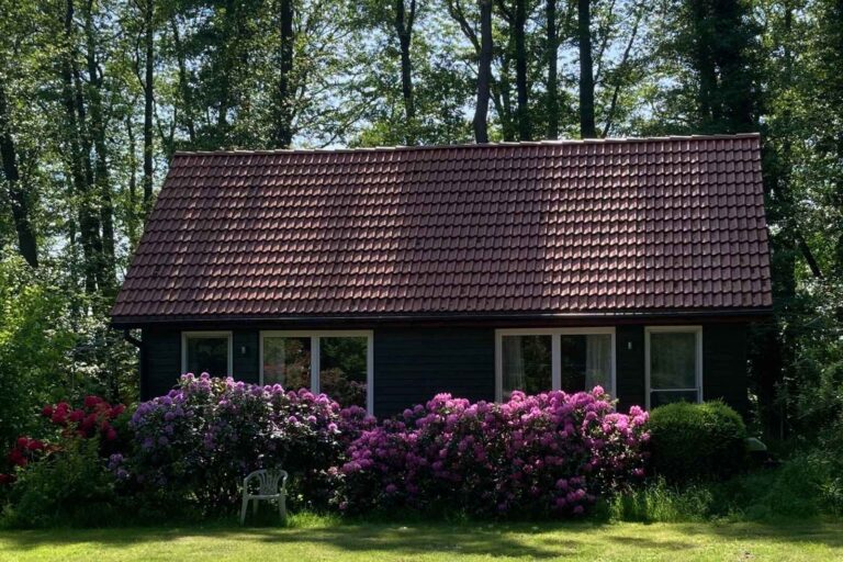 Dunkles Holzgebäude mit rotem Ziegeldach und blühenden Rhododendren, in dem sich die Ferienwohnungen Lichtgrün und Weitblick befinden
